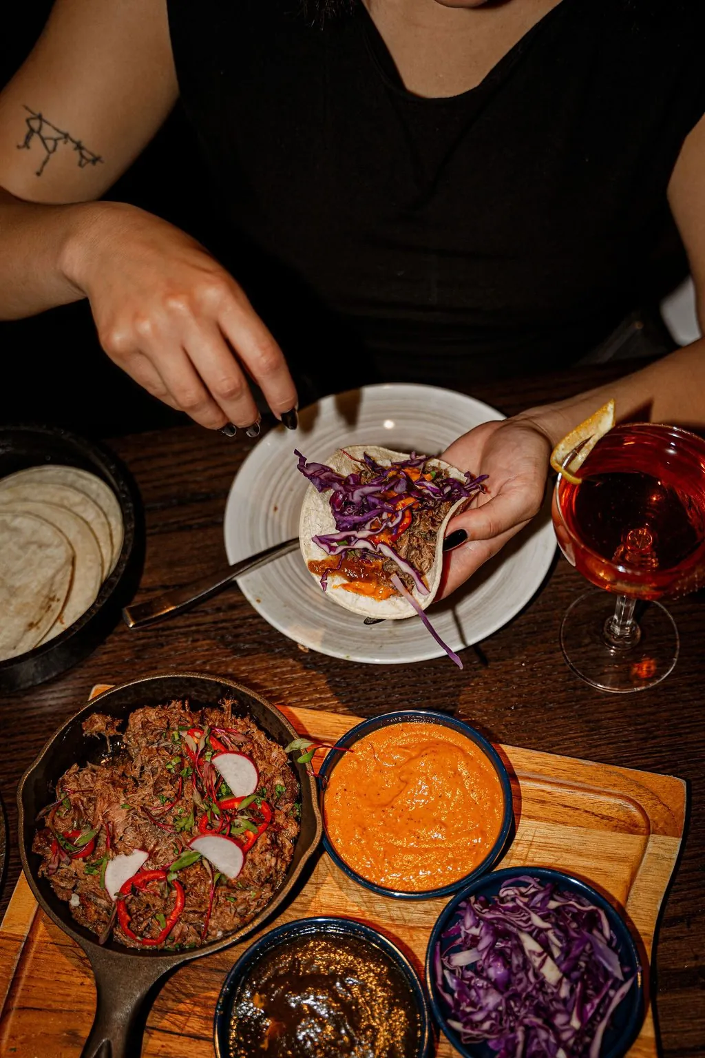 Hands filling a tortilla from a skillet of braised meat, with salsas and cabbage